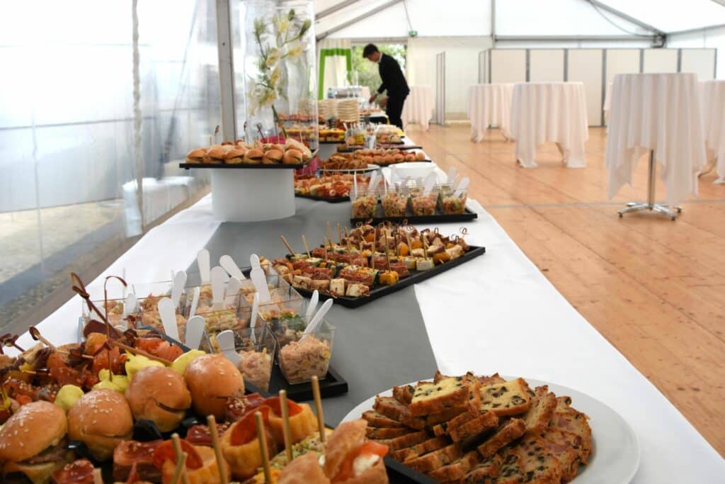 Buffet traiteur avec sélection de mini-burgers, brochettes, verrines et cakes salés. Un serveur en costume prépare la table de réception.